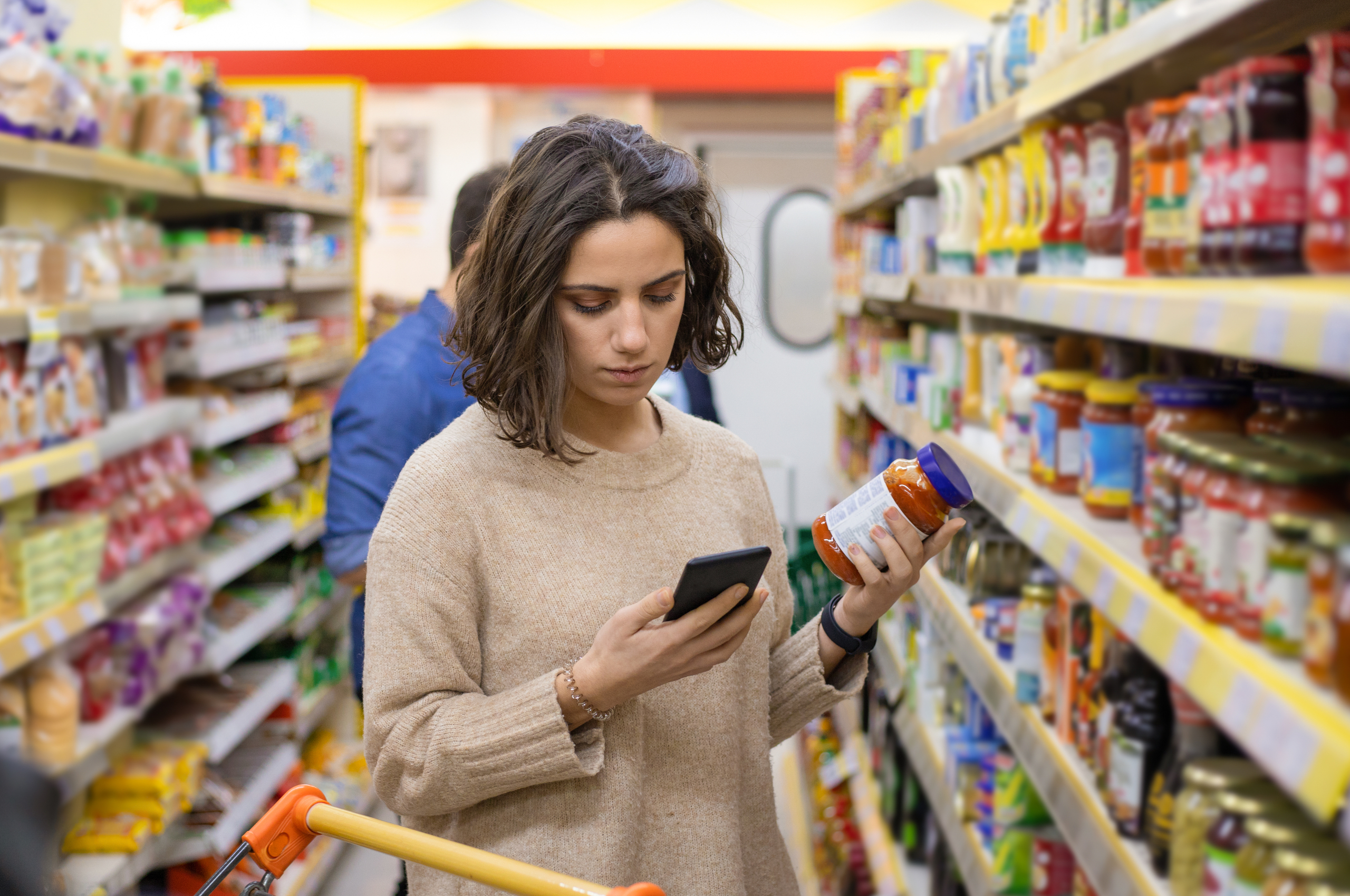 woman looking at a jar in the supermarket while checking her phone