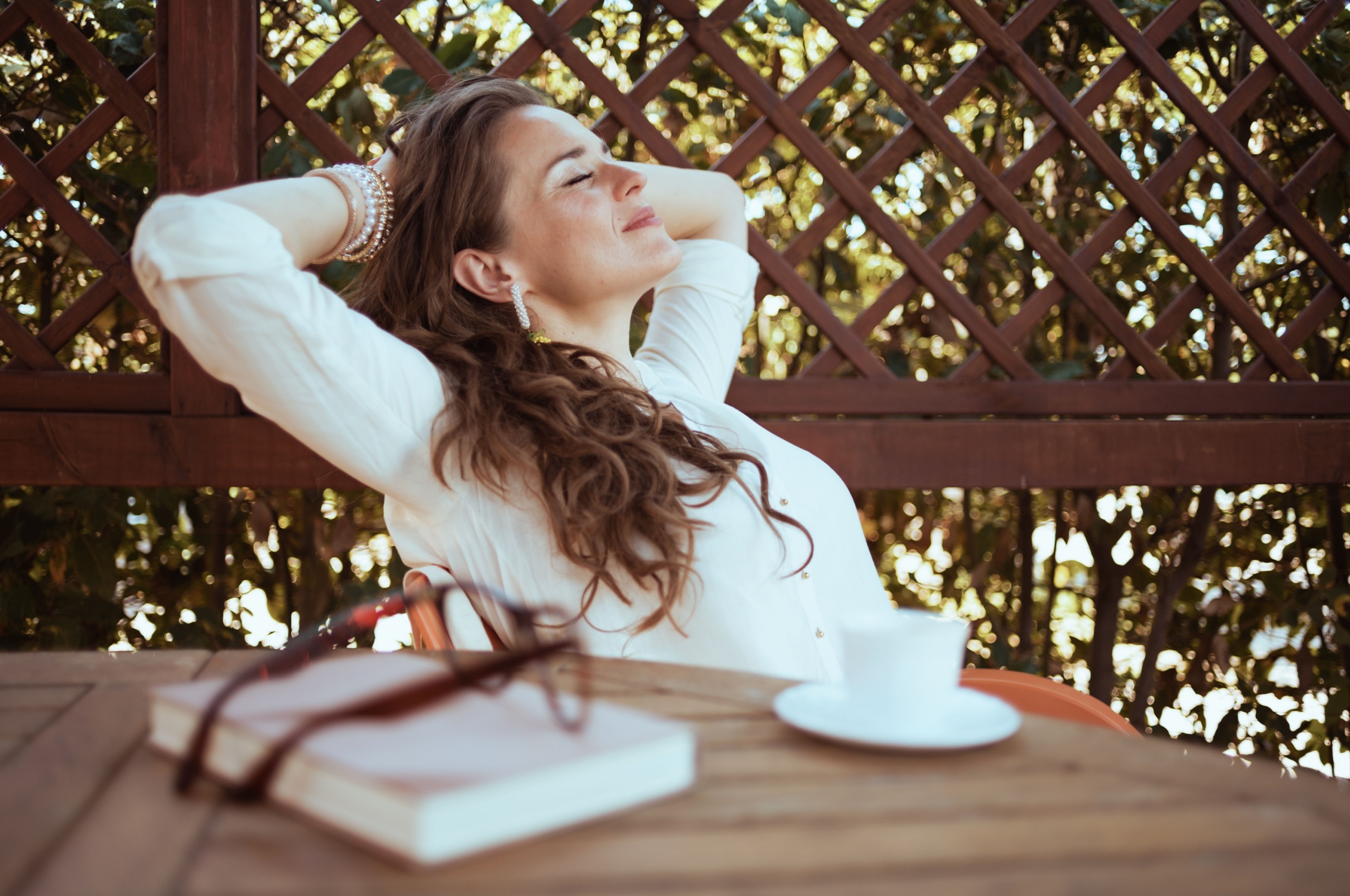 Relaxed middle-aged woman in white shirt with a cup of coffee and a book, sitting outside at a table.